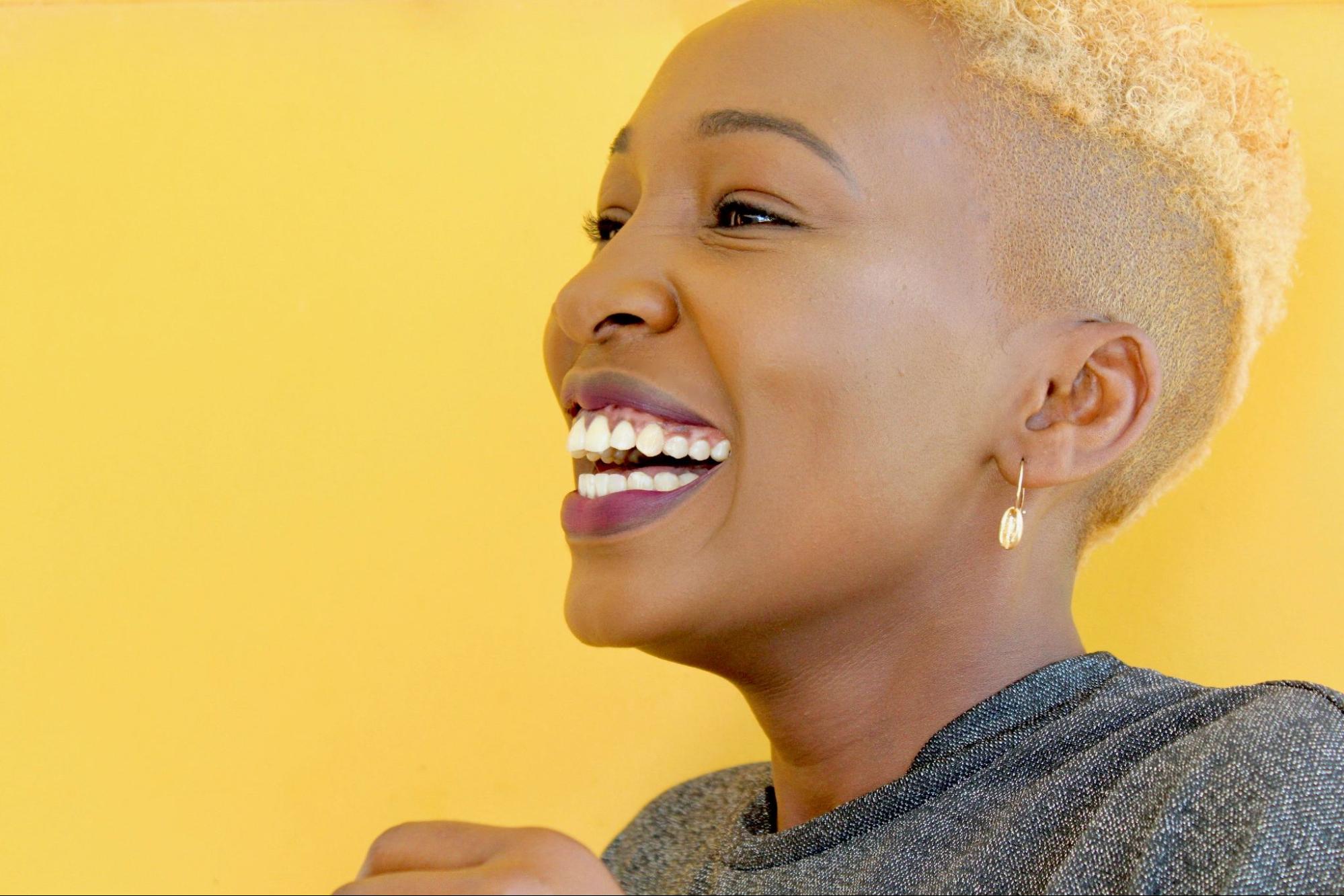 A woman with glowing skin smiles with a yellow background behind her.